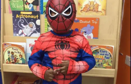 Spiderman child costume standing in front of a bookshelf filled with colorful children's books at Monkey Puzzle Day Nursery Enfield.