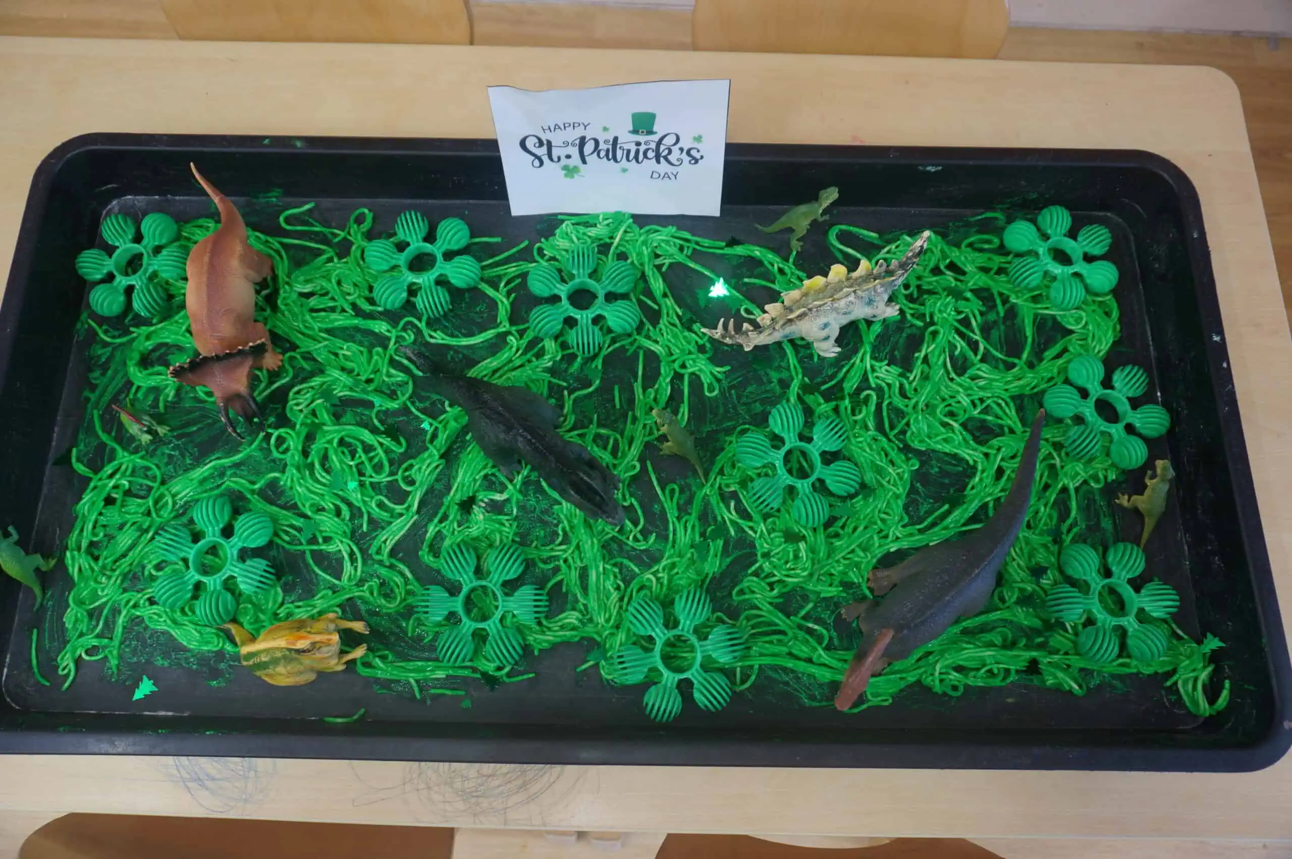 Colorful dinosaur toys and green plastic St. Patrick's Day decorations in a black tray for kids' play, celebrating the holiday at Monkey Puzzle Day Nursery Enfield.
