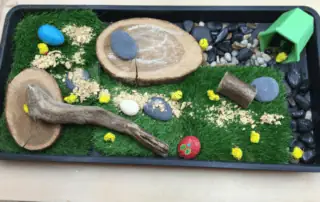 Birch wood slices, small rocks, moss, pebbles, yellow chicks, toy log, green house, and natural sensory materials for children at Monkey Puzzle Day Nursery Enfield.