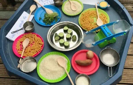 Colorful pretend play food set for children, including pasta, vegetables, and cooking utensils, on a blue tray at Monkey Puzzle Day Nursery Enfield.