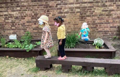 Children playing on a wooden beam in a garden at Monkey Puzzle Day Nursery Enfield, surrounded by flower beds and outdoor learning areas, promoting early childhood development.