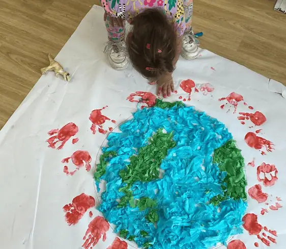 Colorful children's art project with green, blue, and red paint, featuring a child creating a textured Earth craft on a large white paper sheet.
