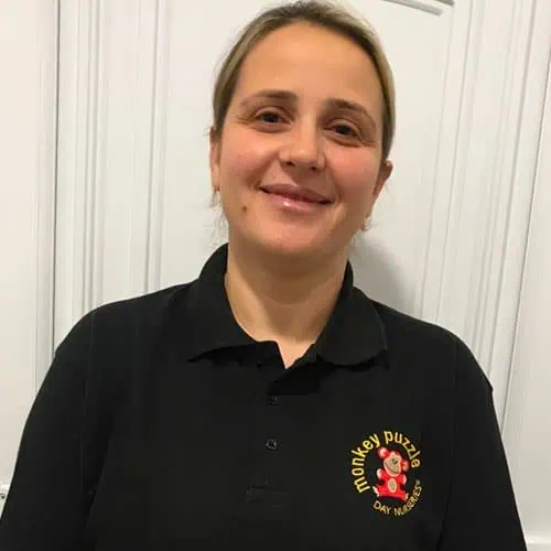 Plump, smiling caregiver in black Monkey Puzzle Day Nursery Enfield uniform standing in front of a white door.