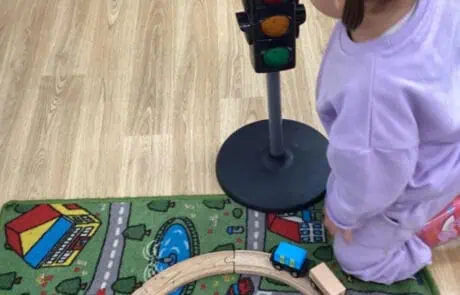 Garden view of a playful child with a toy traffic light, wooden train tracks, colorful train, and city-themed play mat at Monkey Puzzle Day Nursery Enfield, encouraging early childhood development and imaginative learning., Child playing with toy traffic light and train set on colorful play mat at Monkey Puzzle Day Nursery Enfield, promoting early childhood education and imaginative play, with focus on fun learning environments.
