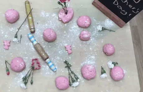 Pink play dough and flower craft items on a table with a "Happy Mother’s Day" chalkboard sign in a preschool setting.
