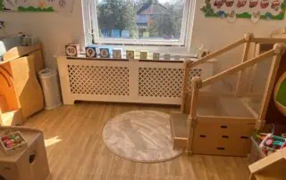 Monkey Puzzle Enfield Environment Wooden indoor play area at Monkey Puzzle Day Nursery Enfield with a view of the outdoors through a window, containing a soft round rug, number blocks, and wooden furniture for children.