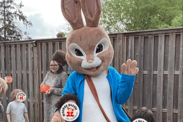 Bunny mascot character at Monkey Puzzle Day Nursery Enfield, outdoor play area, children engaging with staff, friendly educational environment, colorful playground for early childhood development.