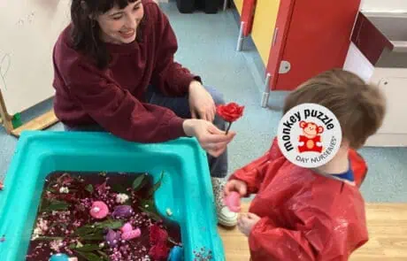 Pink flower with lady holding it, child and teacher in colorful nursery classroom at monkey puzzle day nursery enfield.