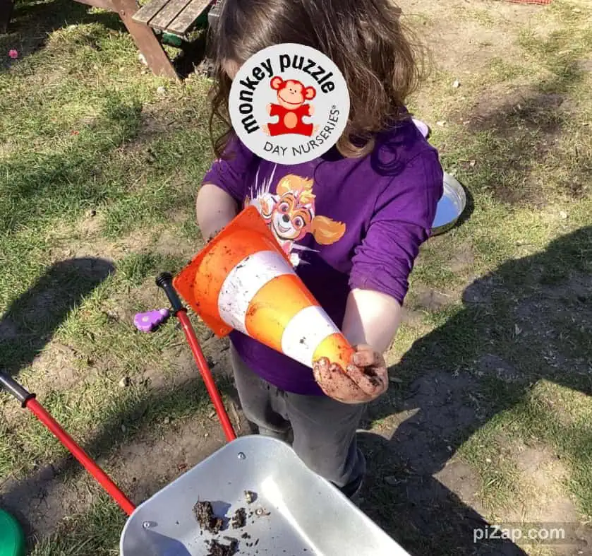 Orange and white traffic cone with mud in child’s hands, outdoors on grassy ground at Monkey Puzzle Day Nursery Enfield.
