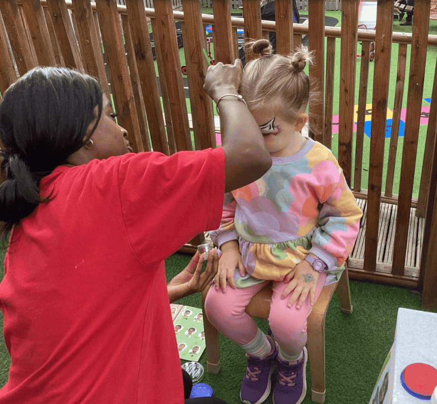 Face painting activity at Monkey Puzzle Day Nursery Enfield, children enjoying creative arts and crafts in an outdoor setting with a staff member assisting; bright, colorful environment.