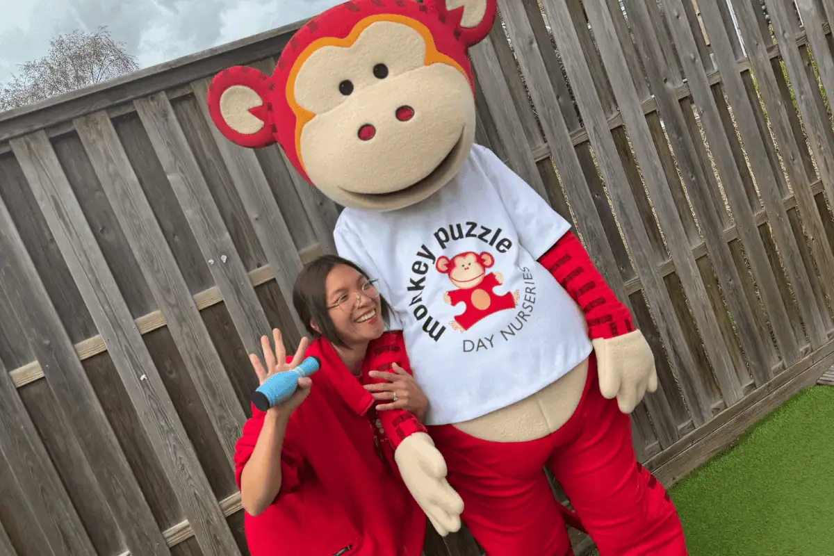 Monkey Puzzle Day Nursery Enfield, cheerful staff with monkey mascot engaging children in playful activities, outdoor garden setting for early childhood development, friendly childcare environment.